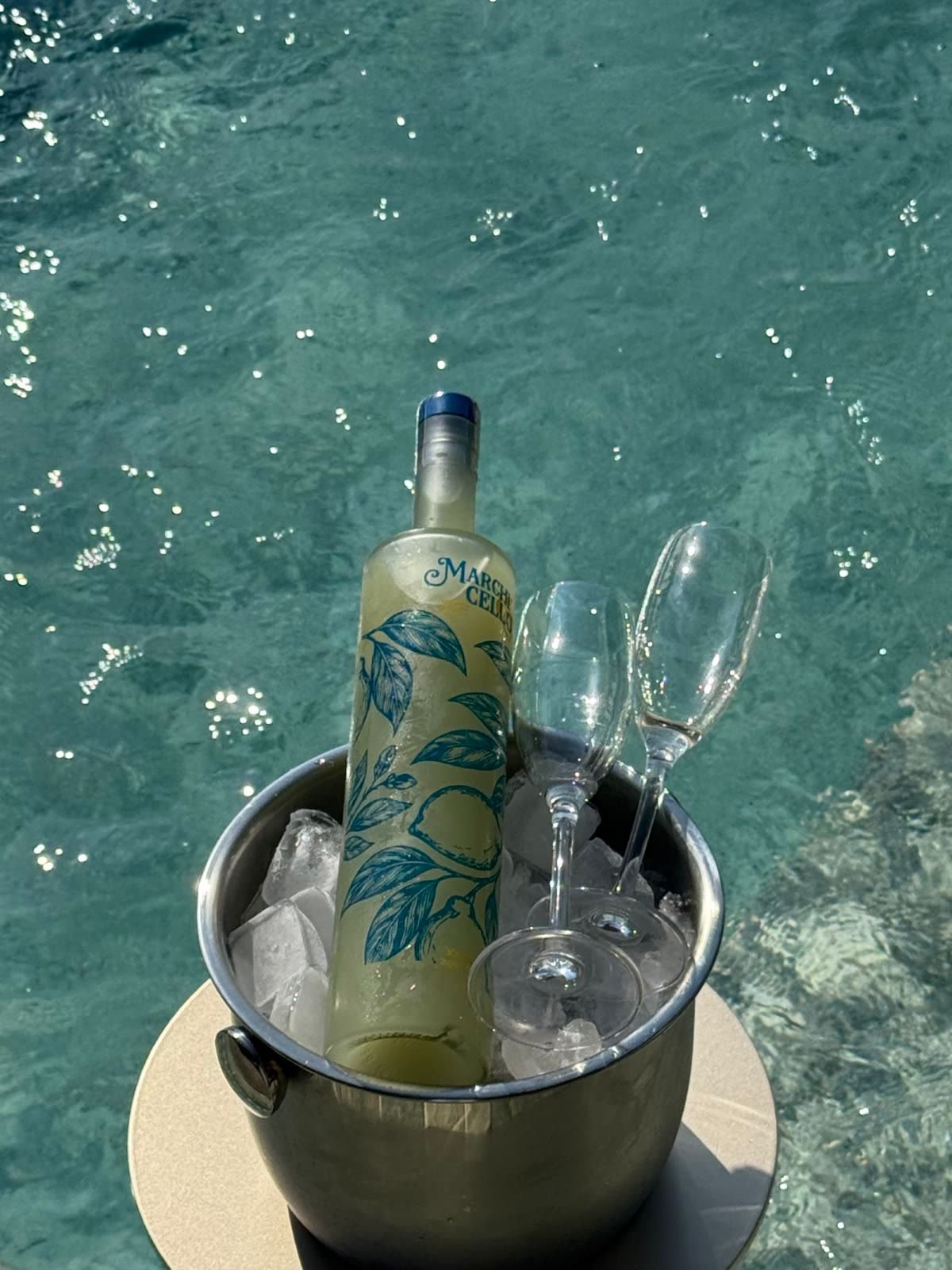 Garrafa de Marchicello e duas taças num balde de gelo de inox sobre a água cintilante da piscina.