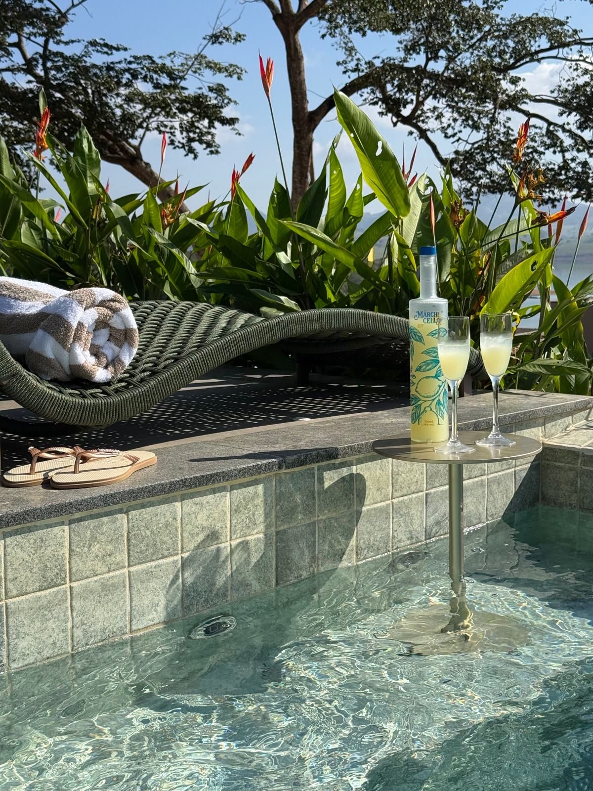 Espreguiçadeira com toalha enrolada ao lado de uma mesinha na piscina, com a garrafa de Marchicello e uma taça, sob uma árvore.
