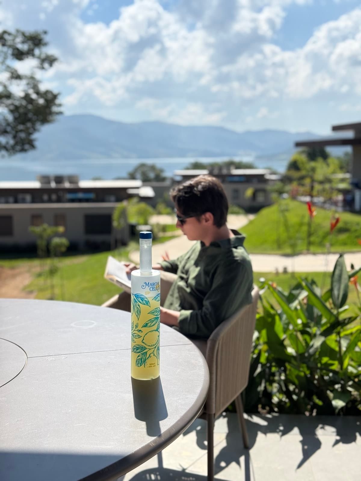 Pessoa de camisa verde lê um livro numa mesa ao ar livre, com a garrafa de Marchicello à frente e o lago e as montanhas ao fundo.